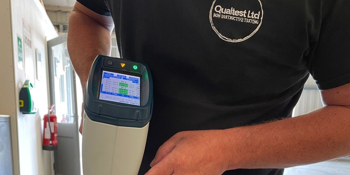 A Qualtest technician in a branded polo holding an XRF analyser in the lab